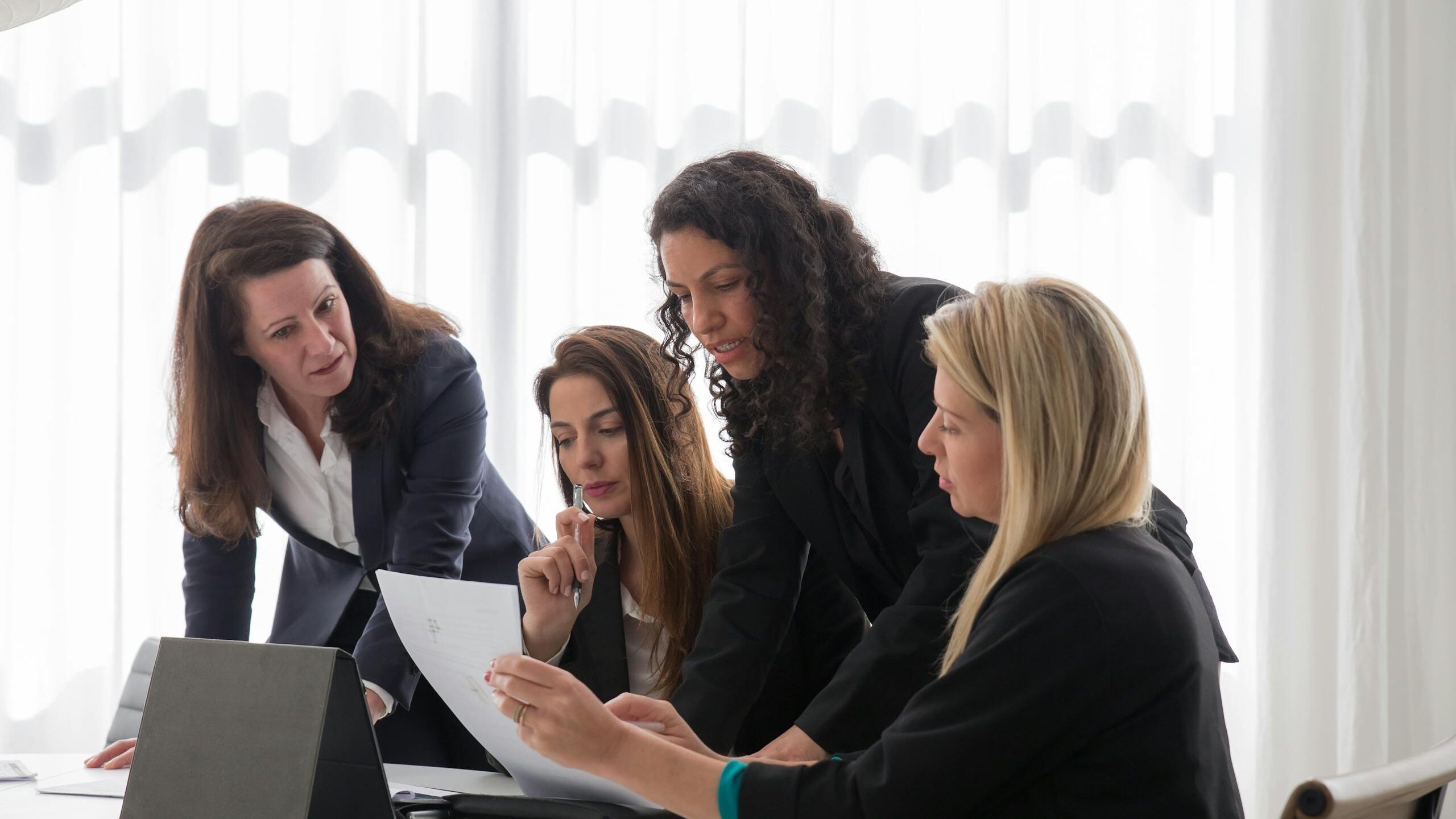 Four businesswomen engaged in discussion over documents in a contemporary office setting.
